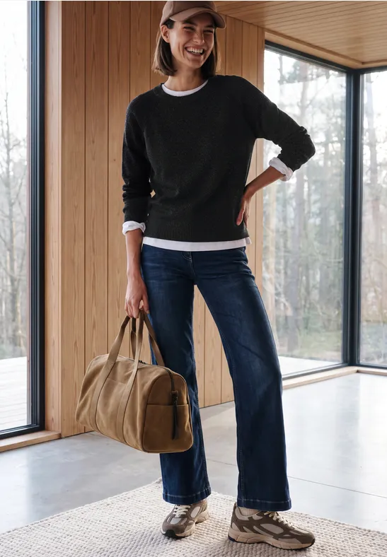 Frau mit schwarzem Pullover, Jeans, Sneakers und Cap hält eine Reisetasche in modernem Holz-Wintergarten.