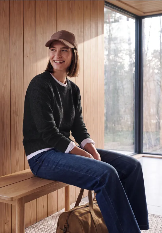 Junge Frau in Cap, Pullover und Jeans sitzt lächelnd auf einer Holzbank am Fenster.
