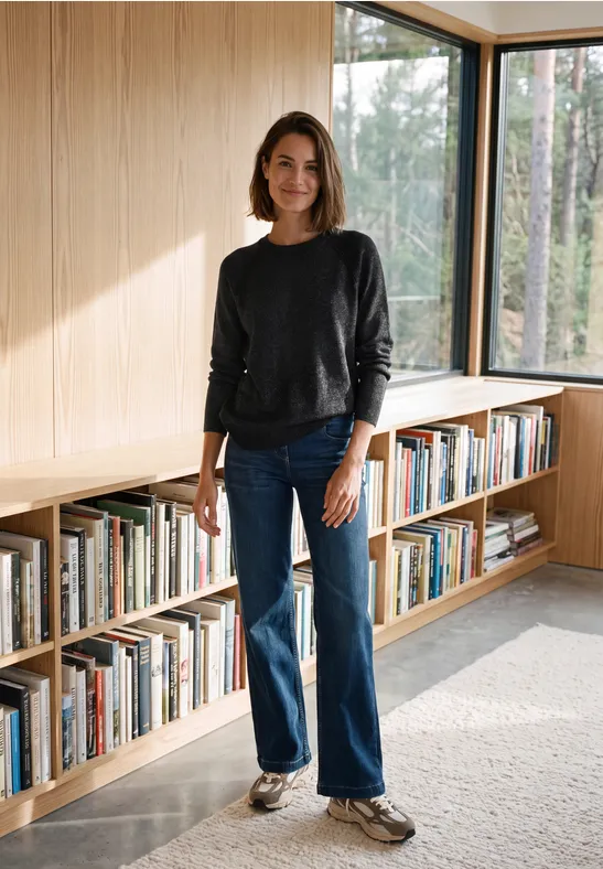 Frau in lässigem Outfit steht vor einem Bücherregal in einem hellen Raum mit großen Fenstern.