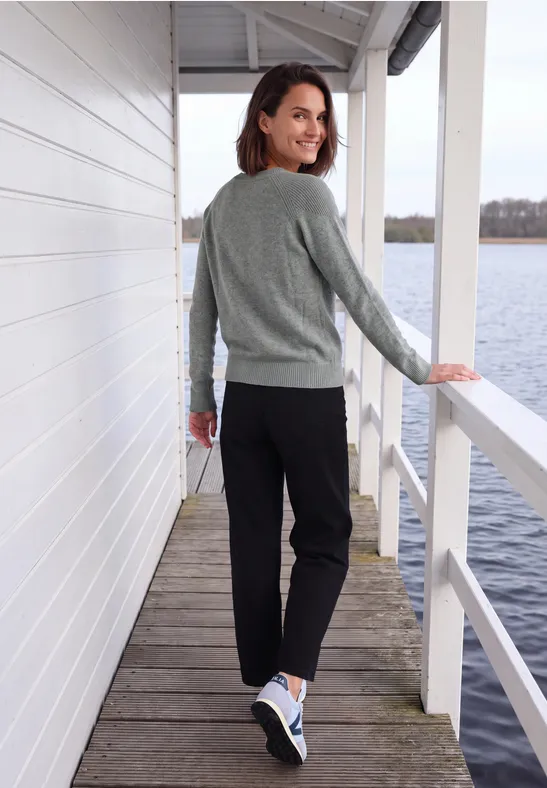 Een vrouw in een grijze trui en zwarte broek loopt over een steiger bij het water en kijkt glimlachend achterom.