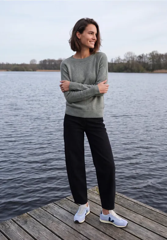 Vrouw staat ontspannen aan het einde van een steiger bij kalm water, gekleed in trui en sneakers.