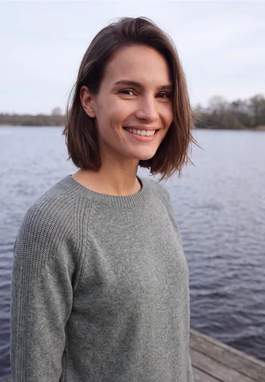 Een jonge vrouw met bruin haar en een grijze trui lacht op een steiger bij het water.