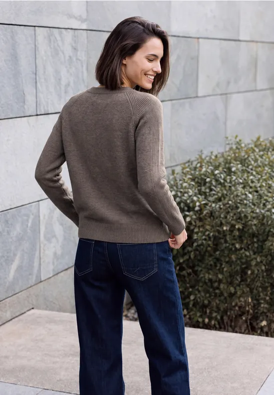 Femme en pull marron et jean devant un mur de pierre gris, sourit en regardant par-dessus son épaule.