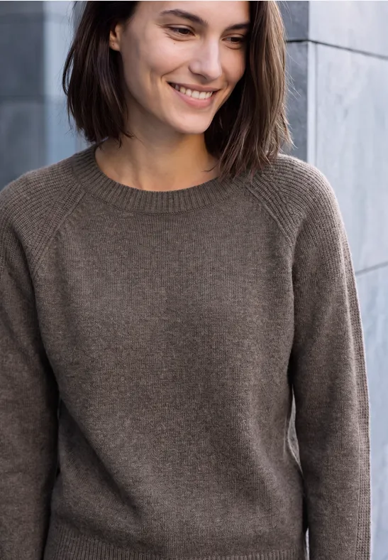 Une femme souriante en pull marron devant un mur de pierre.