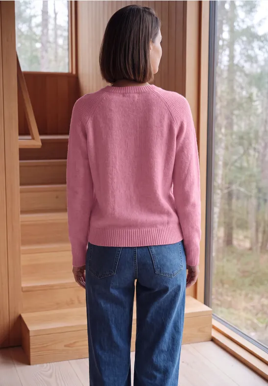 Persoon met roze trui en jeans staat met de rug naar de camera bij grote houten ramen, kijkend naar het bos.