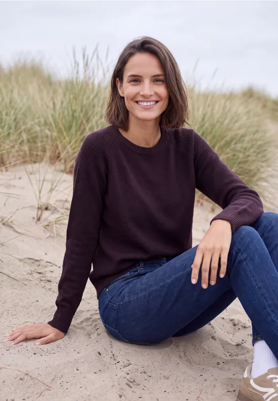 Eine lächelnde Frau sitzt entspannt im Sand vor Dünengras, trägt einen dunklen Pullover und blaue Jeans.