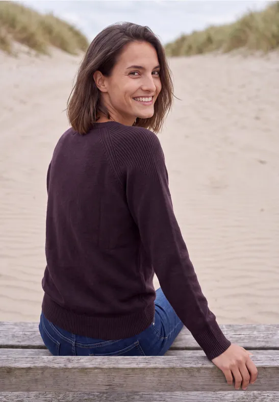 Eine Frau sitzt auf einer Holzbank am Strand, schaut lächelnd über die Schulter zu den Dünen.