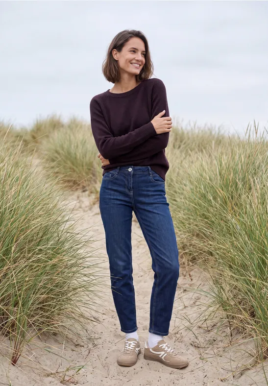 Eine Frau in Jeans und Pullover geht lächelnd zwischen Dünen am Strand spazieren.