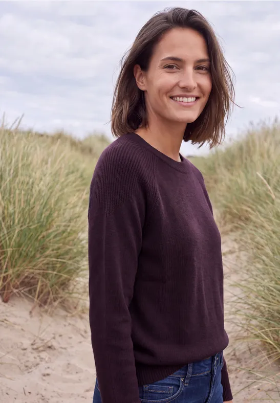 Eine lächelnde Frau mit dunklem Pullover und Jeans, steht vor Dünengras am Strand.
