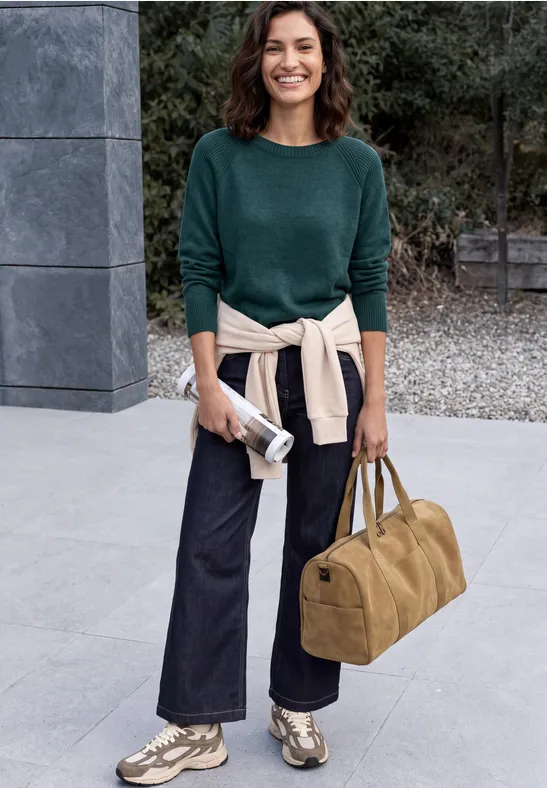 Frau mit grünem Pullover und Jeans steht vor einem Gebäude, trägt Sneakers und hält Tasche und Beutel.