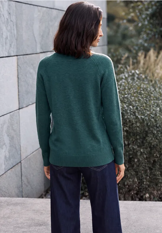 Frau von hinten in grünem Pullover und dunklen Jeans an einer Hauswand und Sträuchern stehend.