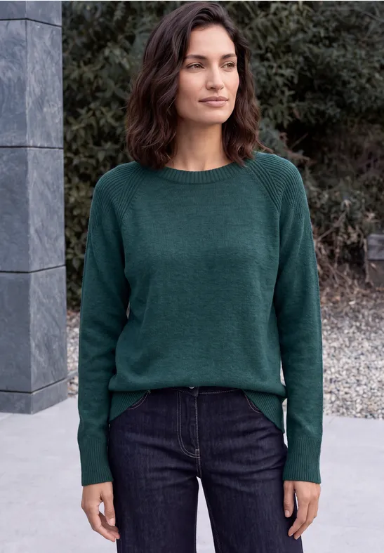 Frau in grünem Pullover und Jeans steht vor Mauer und Büschen im Freien.