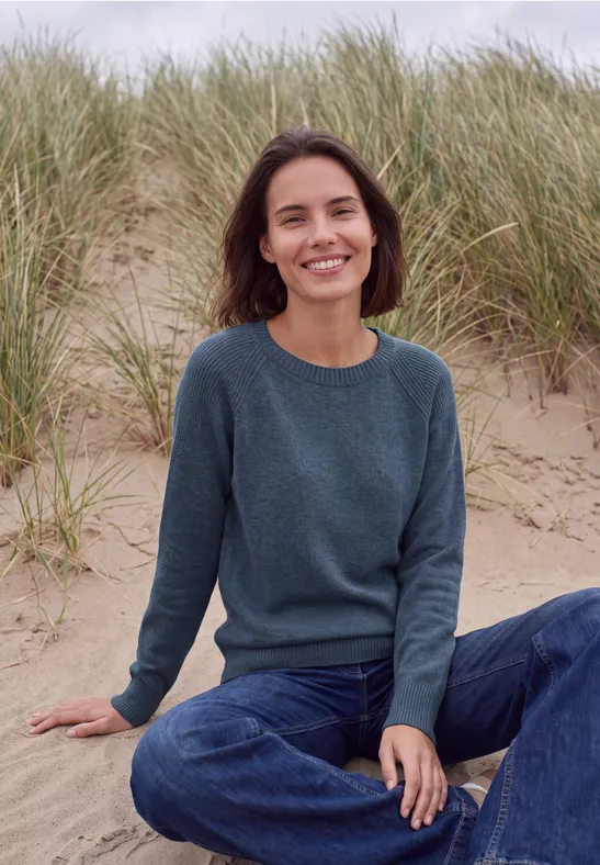 Frau in Jeans und grünem Pullover sitzt lächelnd auf Sanddüne vor Strandgras.