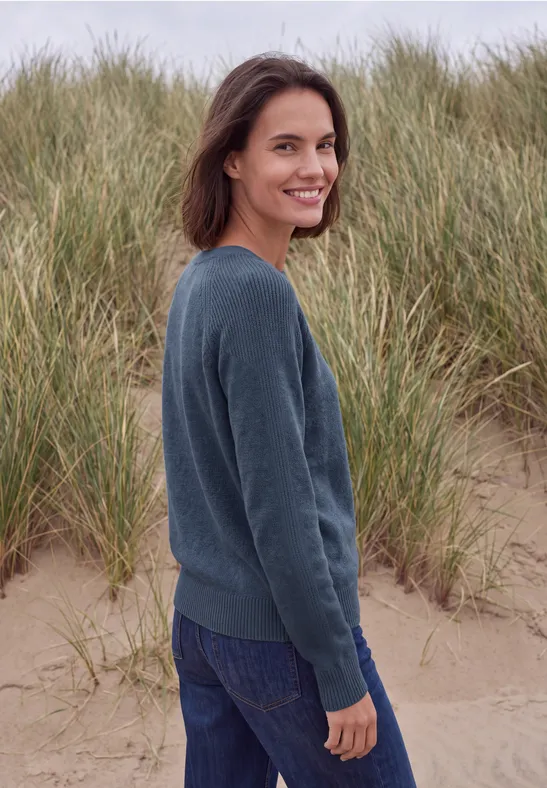 Frau steht lächelnd am Strand vor Dünen, trägt Jeans und Pullover.