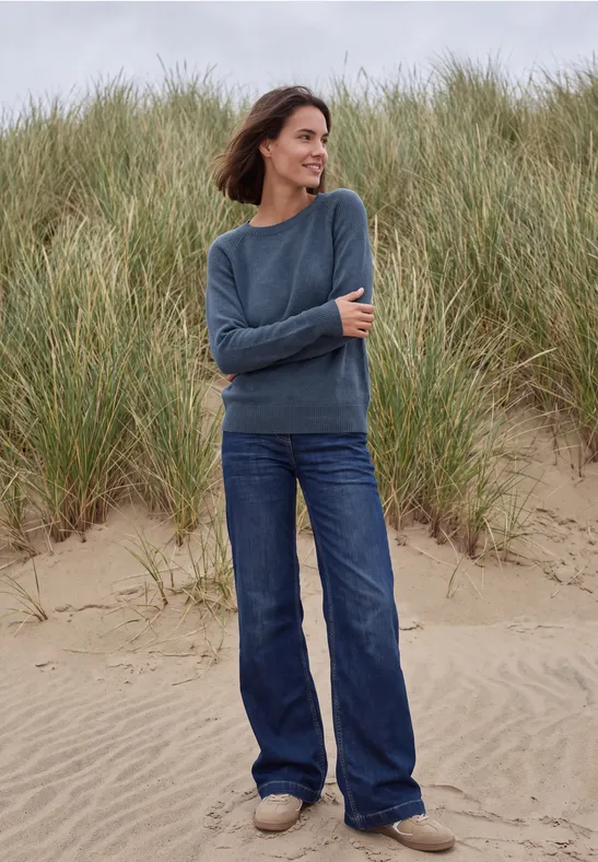 Frau steht am Strand vor Dünen, trägt blauen Pullover und Jeans, lächelnd im Wind.