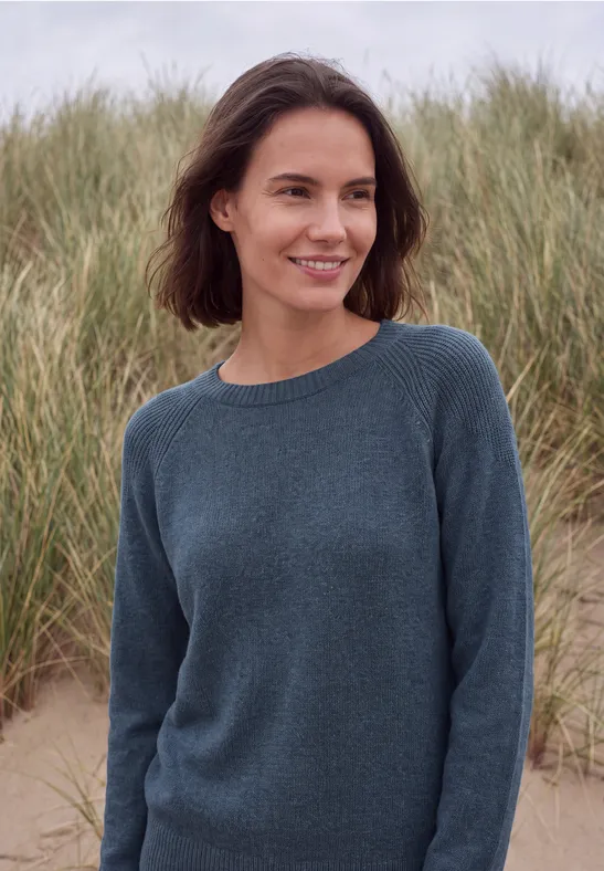 Eine lächelnde Frau in blauem Pullover steht am Strand vor Dünen.