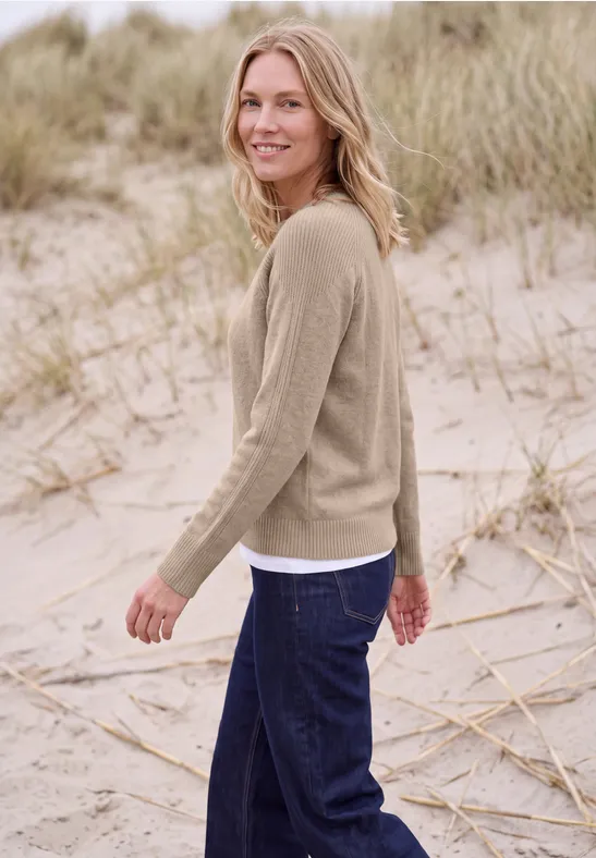 Eine lächelnde Frau in beigem Pullover und dunklen Jeans spaziert am Strand durch Dünen.