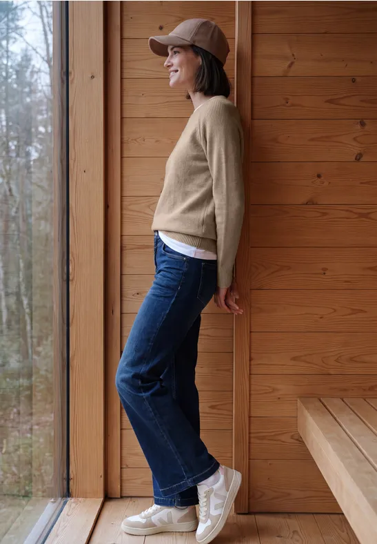 Seitliche Aufnahme einer Frau in Jeans, beigem Pullover und Basecap, die vor einem Holzpaneel lächelnd am Fenster lehnt.