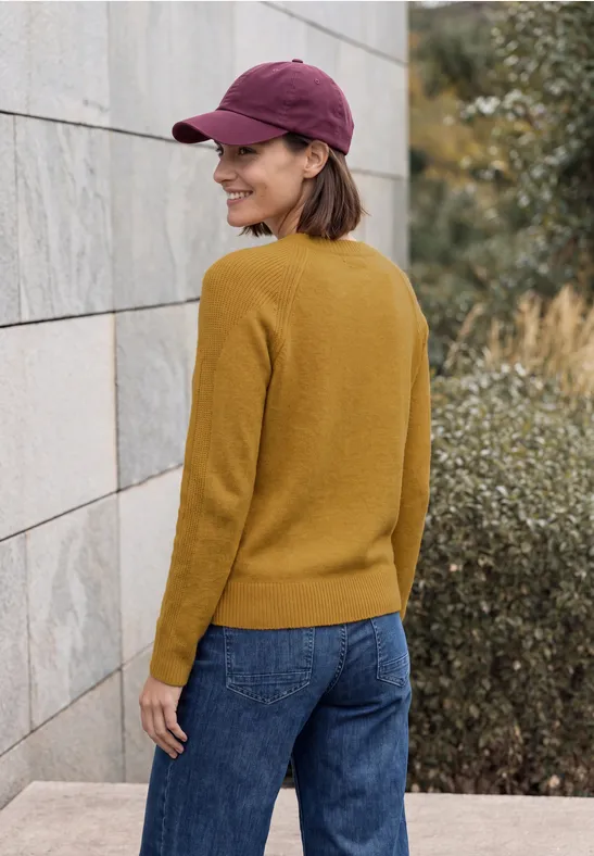 Frau mit gelbem Pullover, Jeans und violettem Cap blickt lächelnd über die Schulter vor Mauer und Grün.