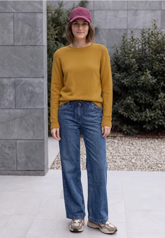 Eine Frau mit senfgelbem Pullover, blauen Jeans, Cap und Turnschuhen steht vor einem modernen Gebäude.