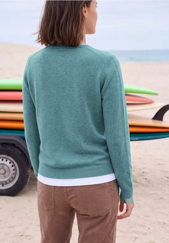 Vrouw in groene trui en bruine broek op het strand voor kleurrijke surfplanken.