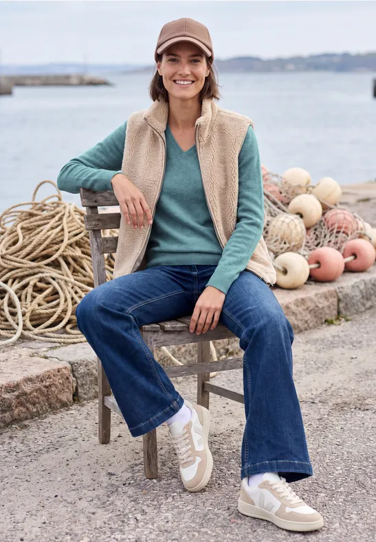Vrolijke vrouw met pet, fleecevest en jeans zit bij de haven tussen touwen en drijvers.