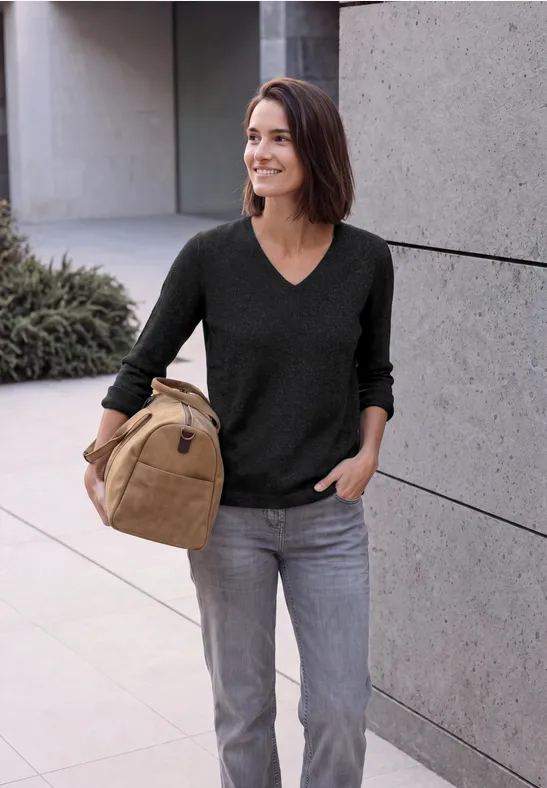 Frau mit dunklem Pullover und grauer Jeans, lächelnd, trägt eine beige Tasche vor moderner Betonwand.
