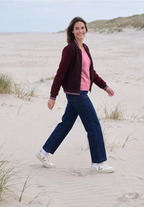 Vrouw in rode jas en spijkerbroek wandelt glimlachend over duinen op het strand.