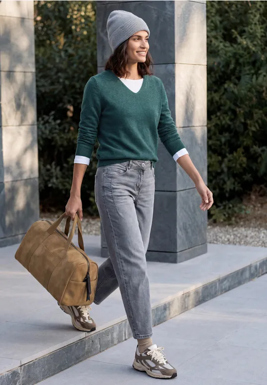 Eine Frau spaziert draußen mit grauer Jeans, grünem Pullover, Mütze und Tasche.