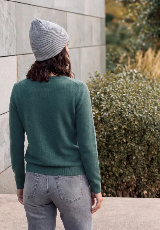 Frau mit grauem Beanie und grünem Pullover steht mit dem Rücken zur Kamera vor einer Mauer und Büschen.