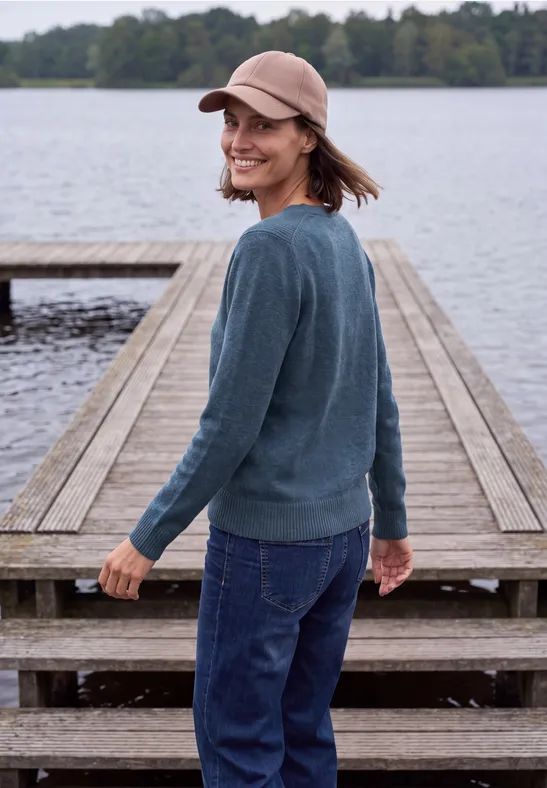 Eine lächelnde Frau mit Mütze und Pullover steht auf einem Steg am ruhigen See.