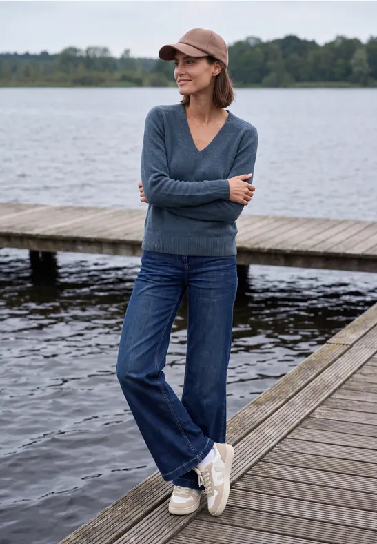 Frau mit Jeans, Pullover und Cap steht lächelnd auf einem Holzsteg am See.
