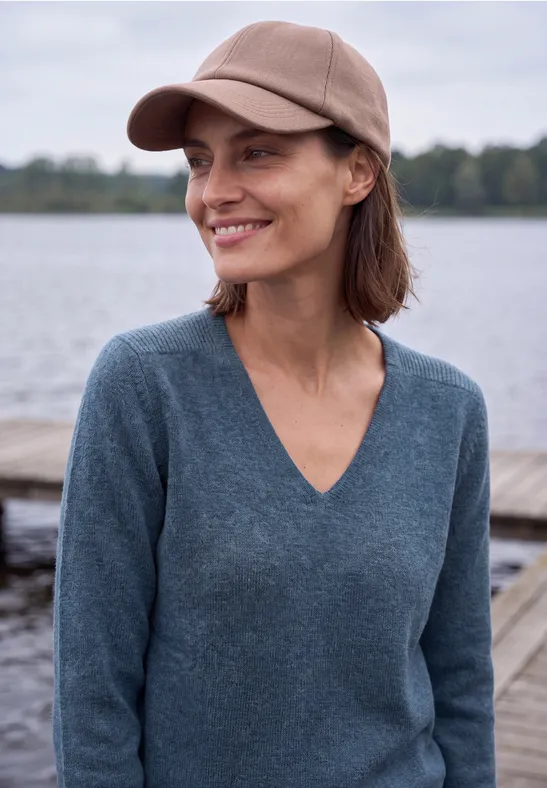 Eine lächelnde Frau mit Baseballcap und Pullover steht am Seeufer.