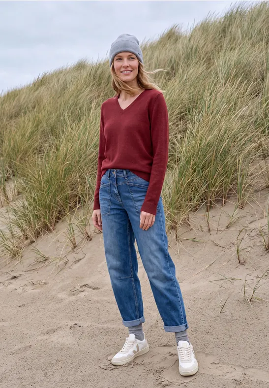Een glimlachende vrouw met grijze muts, rode trui, blauwe jeans en witte schoenen op een zandduin.