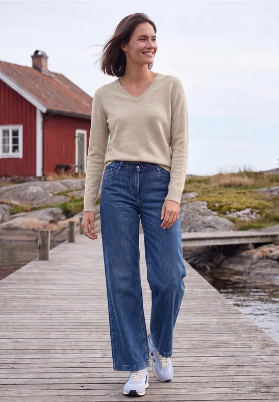 Eine Frau in Jeans, beigem Pullover und weißen Sneakers spaziert auf einem Holzsteg mit rotem Haus im Hintergrund.