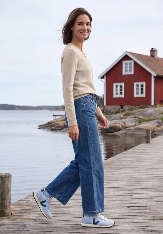 Frau in Jeans und Pullover steht lächelnd auf einem Steg vor einem roten Haus am Wasser.