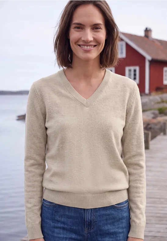 Eine lächelnde Frau in beigem V-Ausschnittpullover und Jeans steht vor einem roten Haus am See.