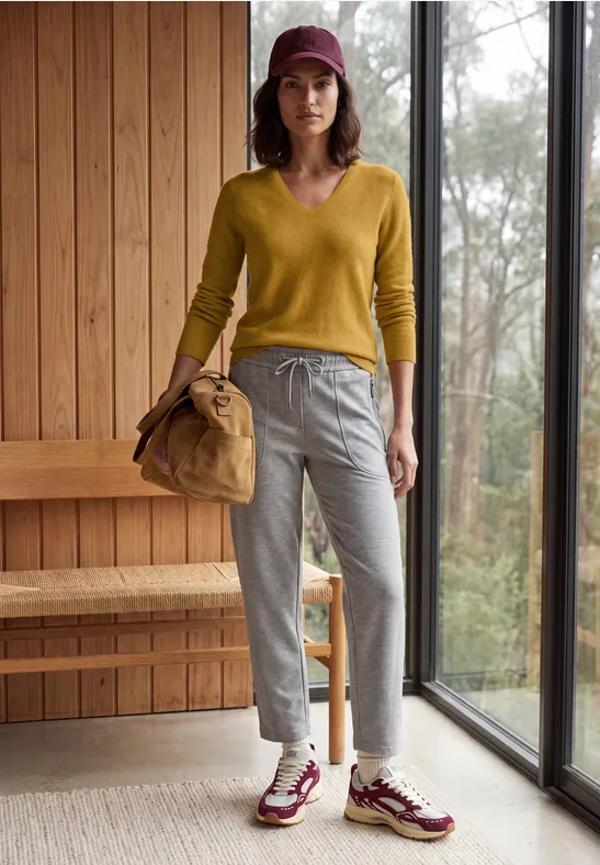 Frau in gelbem Pullover, grauen Hosen, bordeauxfarbenem Hut und Sportschuhen, mit Tasche neben Fenster.
