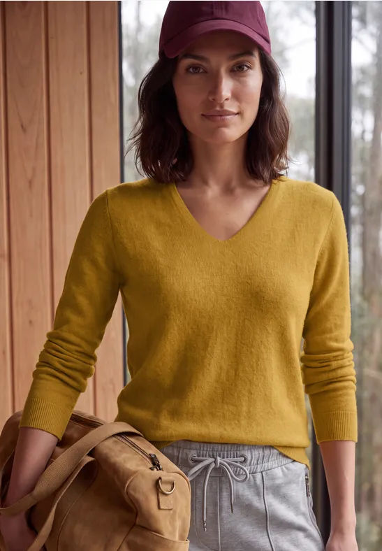 Eine Frau in gelbem Pullover, grauen Jogginghosen und roter Kappe hält eine beige Tasche und steht drinnen.
