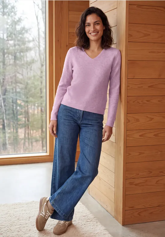 Eine Frau in rosa Pullover und Jeans steht lächelnd vor einem Holzfenster mit Blick ins Freie.