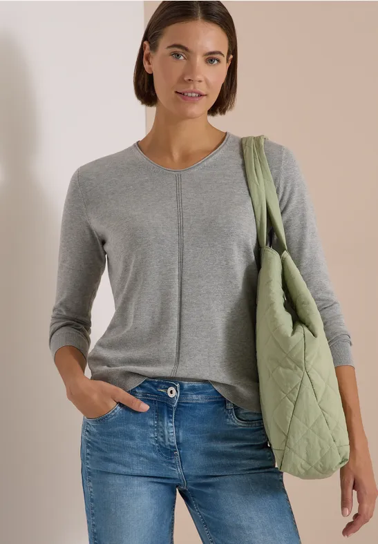 Eine Frau trägt einen grauen Strickpullover, blaue Jeans und eine olivgrüne Stepptasche über der Schulter.