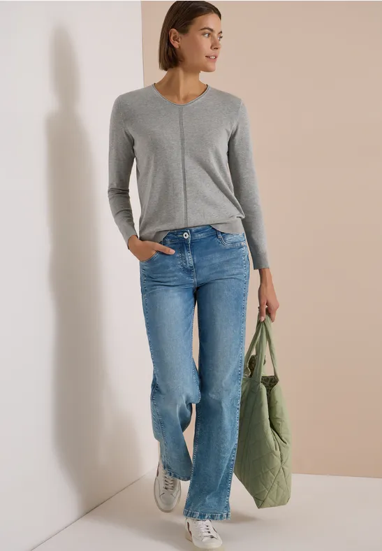 Frau in grauem V-Pullover, lässigen blauen Jeans, weißen Sneakern und grünem Shopper vor hellem Hintergrund.