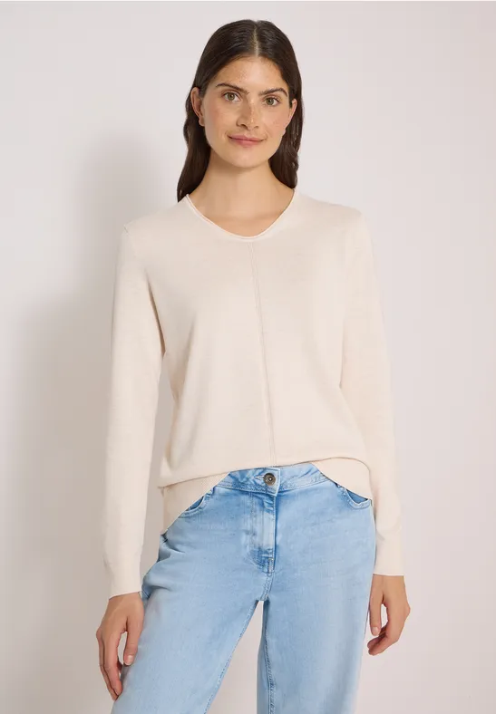 Frau mit hellem Pullover und heller Jeans in lässiger Pose vor neutralem Hintergrund.