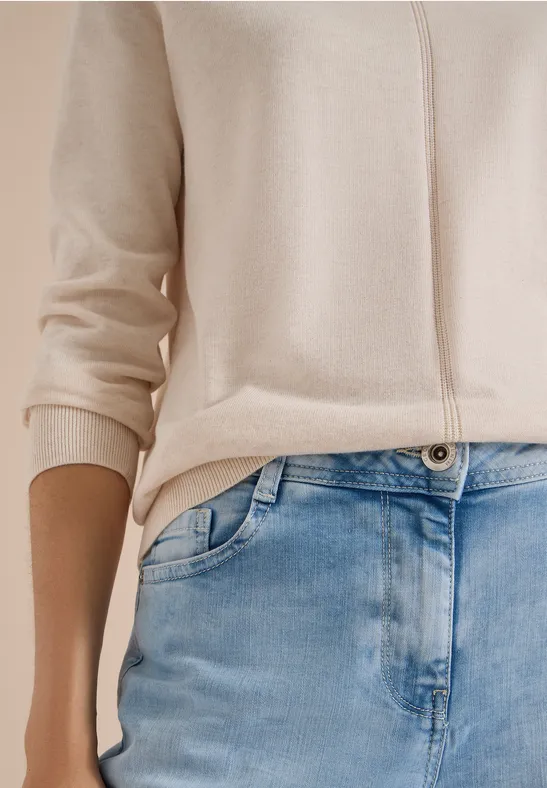 Beige Cardigan und blaue Jeans an einer stehenden Person, Fokus auf Taille und Oberkörper.