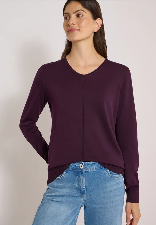 Eine Frau trägt einen dunkelvioletten V-Ausschnitt-Pullover und blaue Jeans vor hellem Hintergrund.