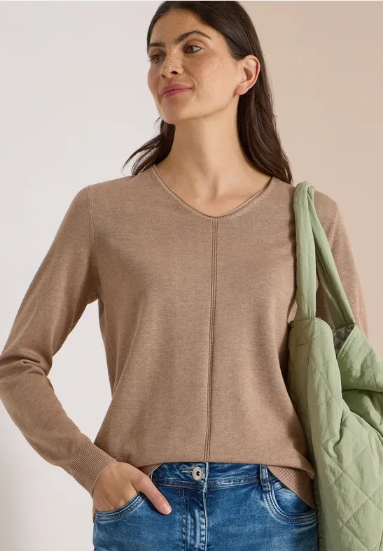 Frau mit beige V-Pullover und blauer Jeans, grüne Steppjacke lässig über der Schulter.