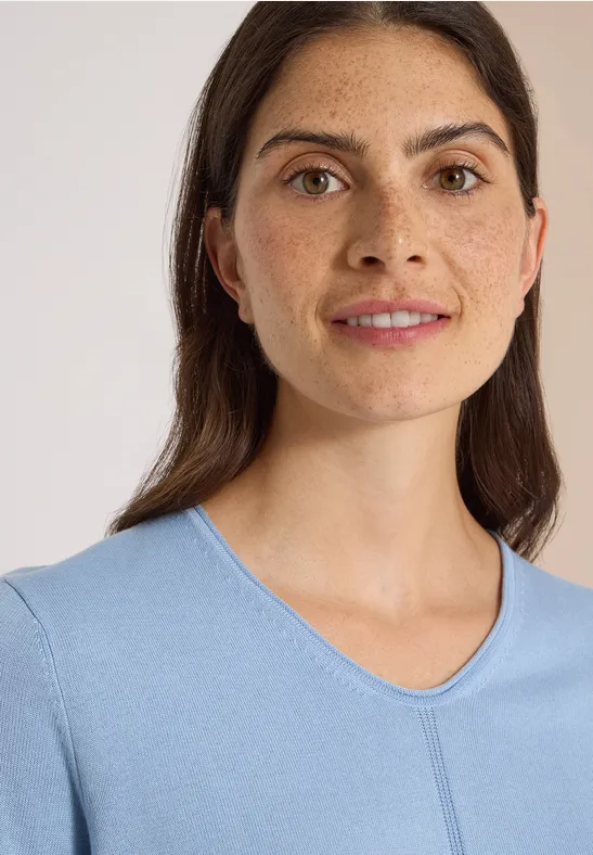 Porträt einer lächelnden Frau mit dunklem Haar in einem hellblauen Shirt vor hellem Hintergrund.