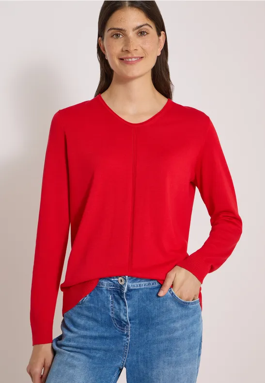 Frau in rotem V-Ausschnitt-Pullover und blauen Jeans, eine Hand in der Tasche.