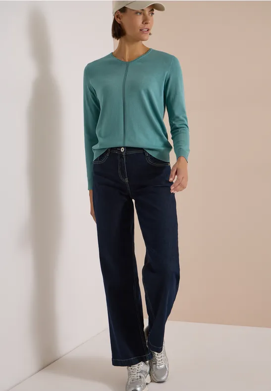 Eine Frau in grüner Strickjacke, Jeans und Sneakern vor hellem Hintergrund.