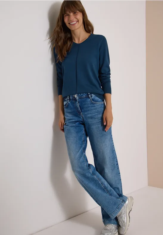 Eine lächelnde Frau in Jeans, Sneakern und blauem Oberteil lehnt an einer Wand.